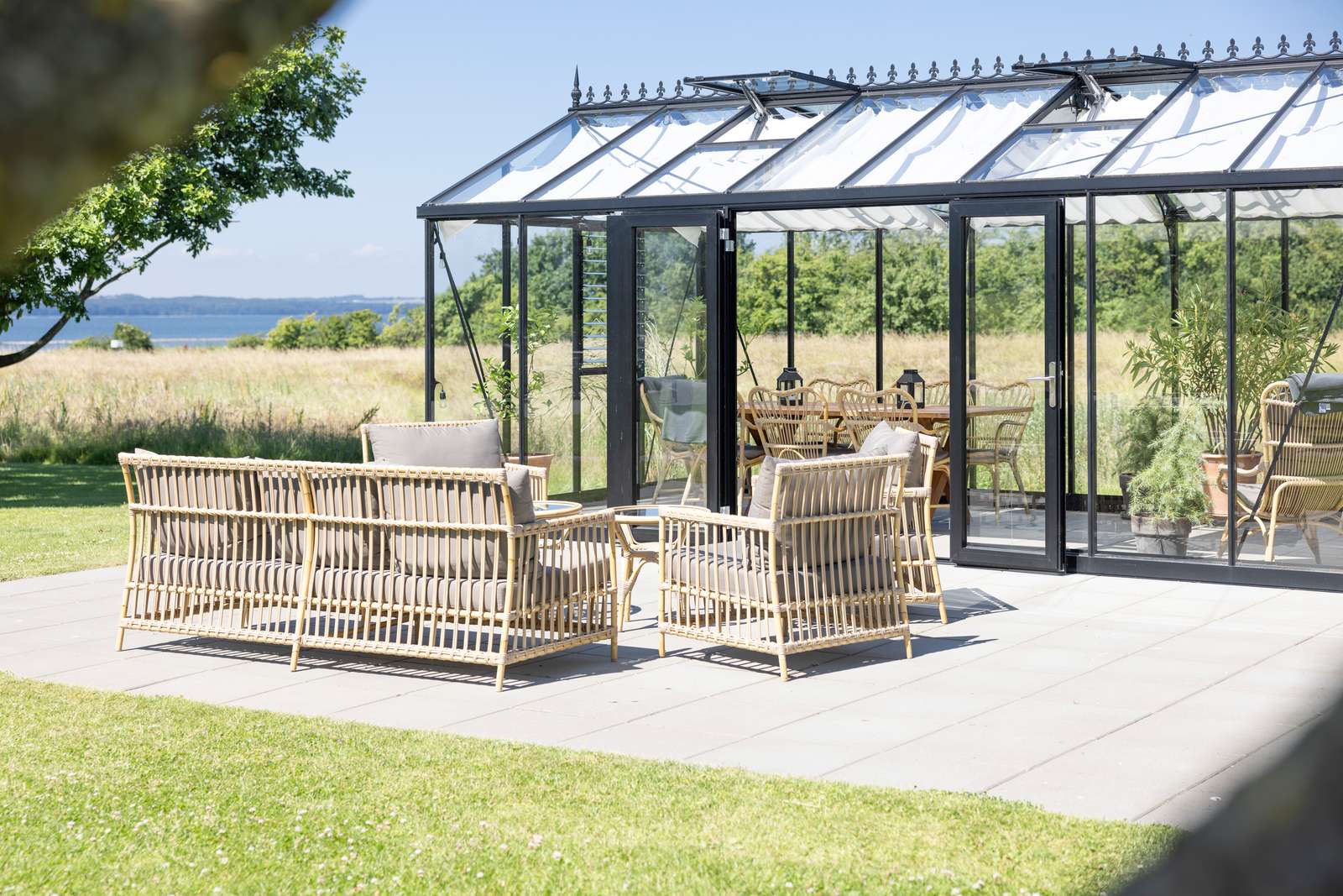 Væksthus eller orangeri, der er indrettet med kurvemøbler, ses udefra med hav i baggrunden 