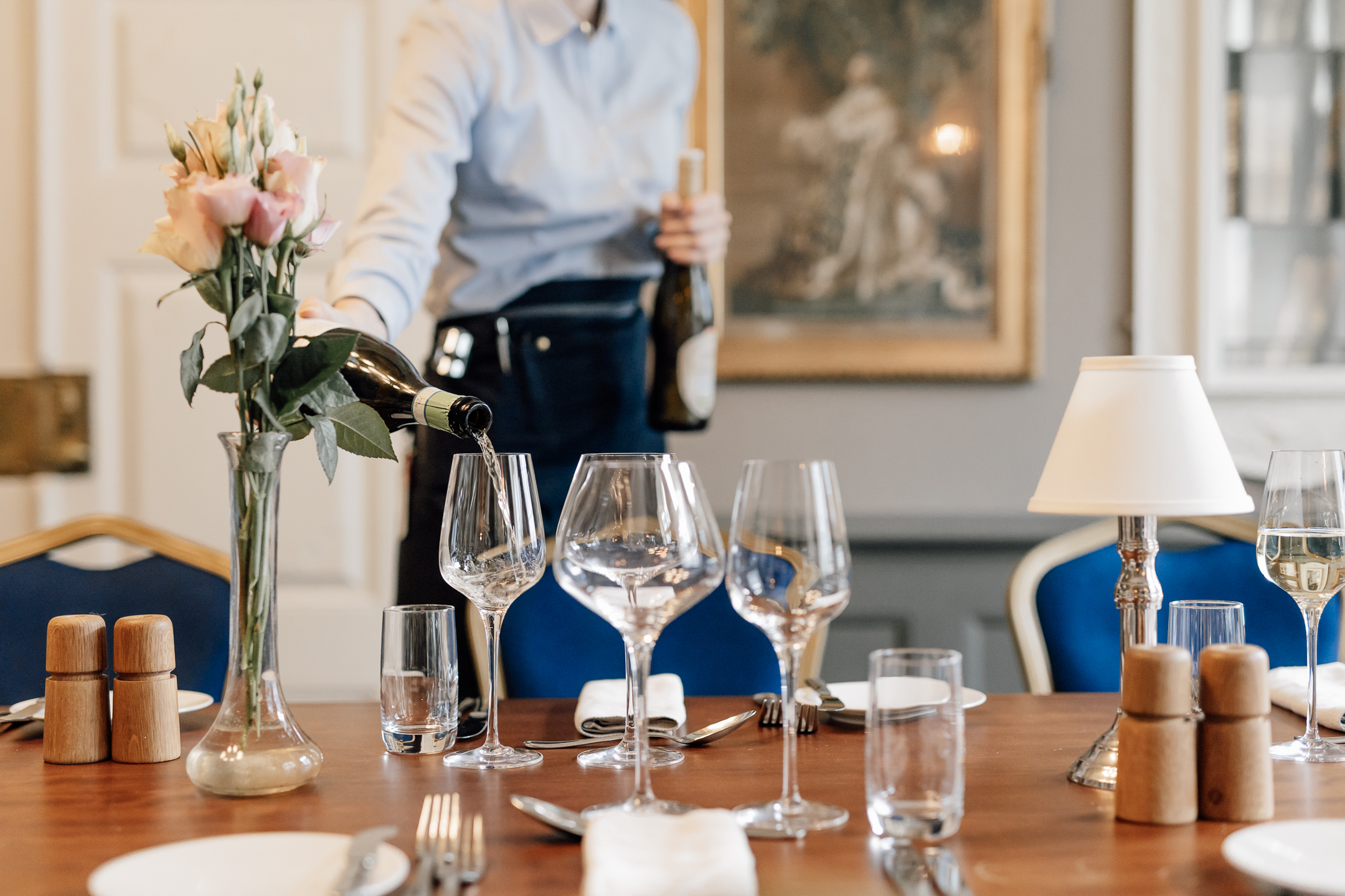 En tjener i blå skjorte hælder vin op i et glas på et flot dækket bord 