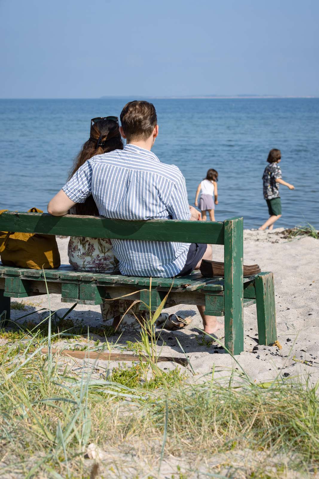 Par sidder på en bænk på Hou Strand, mens to børn leger