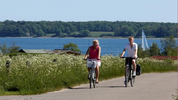 Two cyclists ride along a road with water in the background